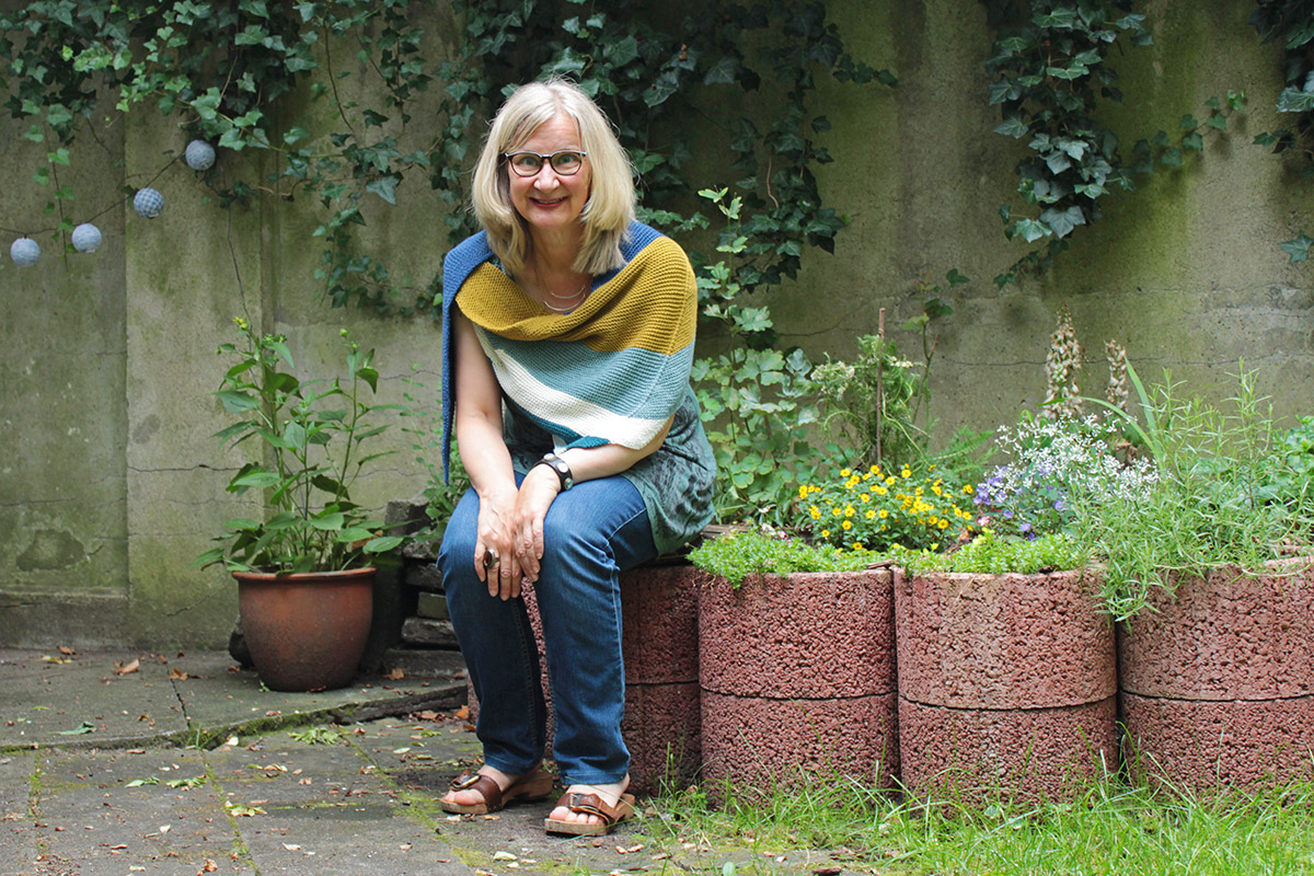 Eine Frau sitzt auf einer Gartenmauer und trägt ein selbstgestricktes Schultertuch aus Wolle