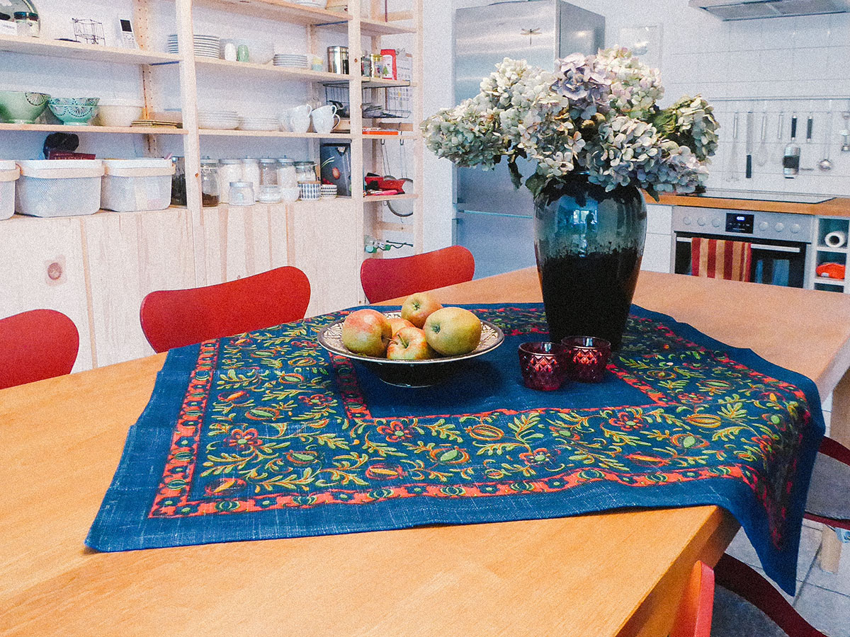 Ein bunt bedruckte Decke liegt auf einem Tisch darauf eine Schale mit Äpfeln und eine Vase mit getrockneten Hortensien