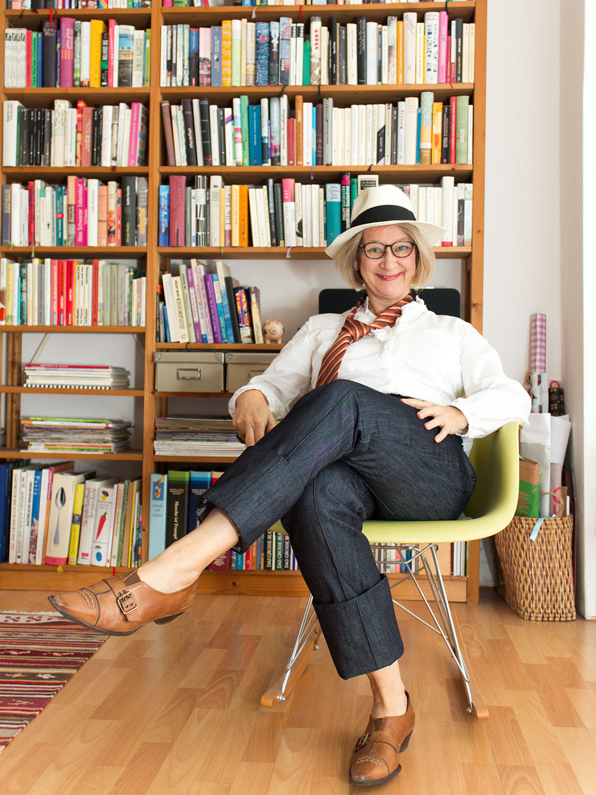 Frau in Jeanshose, weißem Hemd und Krawatte sitzt in einem Sessel vor einem Bücherregal