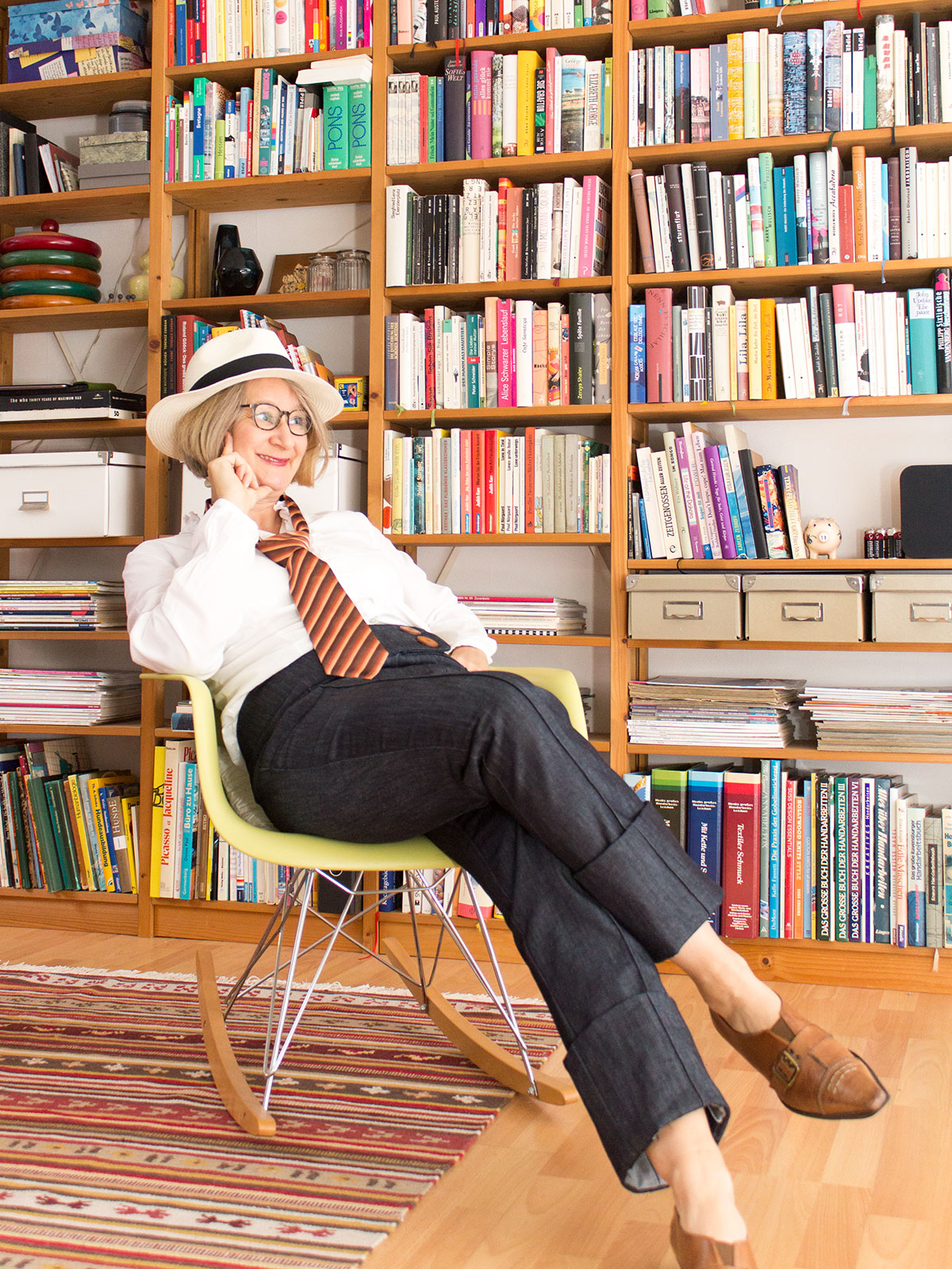 Frau in Jeanshose, weißem Hemd und Krawatte sitzt in einem Sessel vor einem Bücherregal
