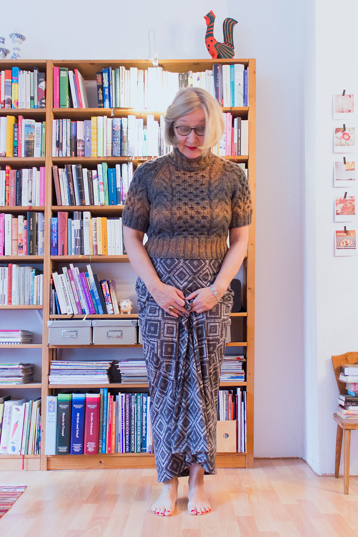 Eine Frau in einem handgestrickten Rollkragenpullover mit kurzen Ärmeln und einem langen Kleid steht vor einem Bücherregal