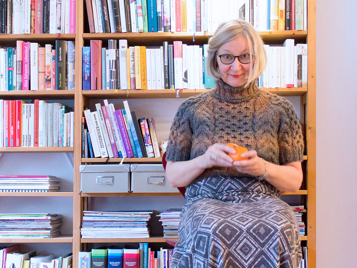 Eine Frau in einem handgestrickten Rollkragenpullover mit kurzen Ärmeln und einem langen Kleid sitzt auf einem Stuhl vor einem Bücherregal