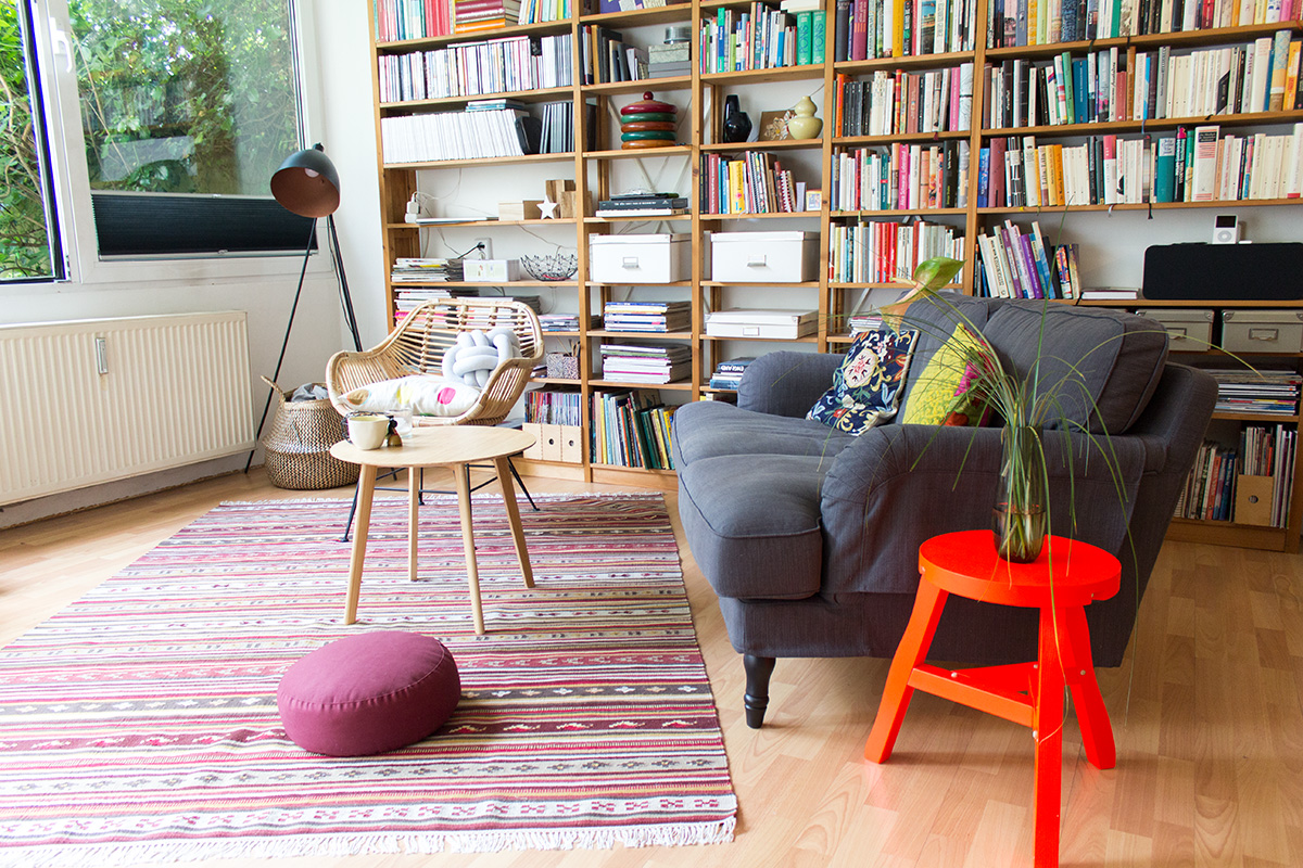 Wohnzimmersituation: Sofa, Coffetable, Rattansuhl, bunte Kissen, Bücherregal, IKEA Teppich