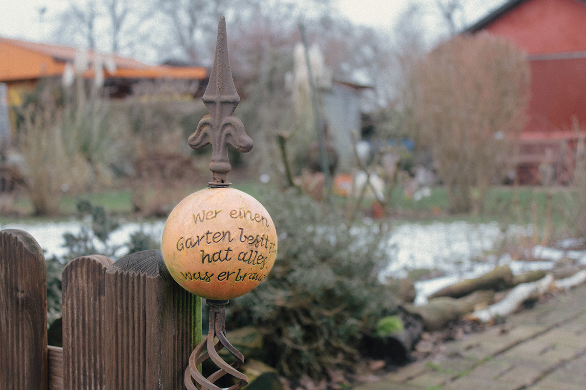 Ausschnitt aus einem Schrebergarten: ein gusseiserner Stab mit einer angebrachten Tonkugel darauf der Spruch "Wer einen Garten besitzt, hat alles, was er braucht."