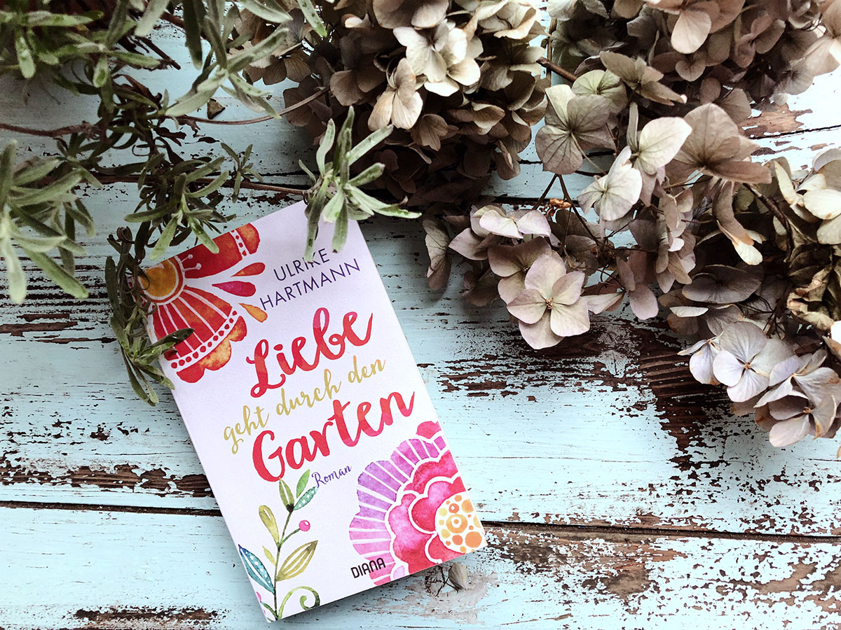 Ein Buch mit dem Titel "Liebe geht durch den Garten" liegt auf einem türkisfarbenen Gartentisch umringt von Lavendel und getrockneten Hortensien