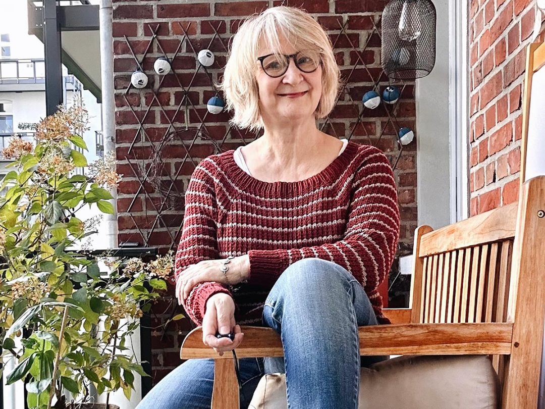 Eine blonde Frau in einem Ringelpullover sitzt lässig auf einer Bank und lächelt in die Kamera
