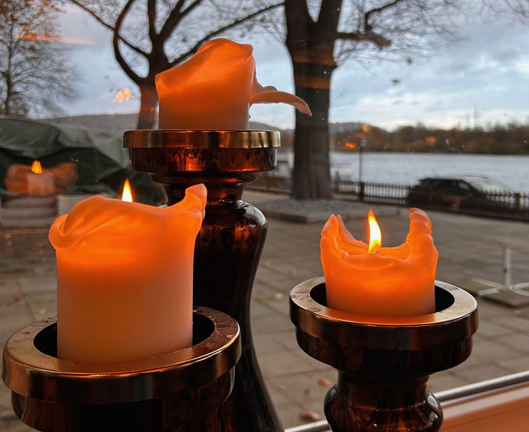 Drei brennende, dicke Kerzen vor einem Fenster dahinter eine Novemberlandschaft und ein See