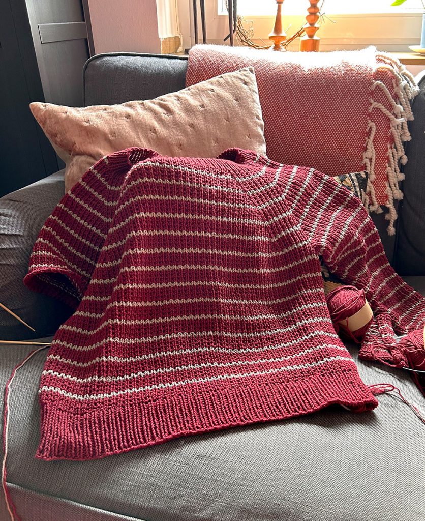 Ein halbfertiges Strickstück in Baumwolle liegt auf einem Sofa mit Kissen